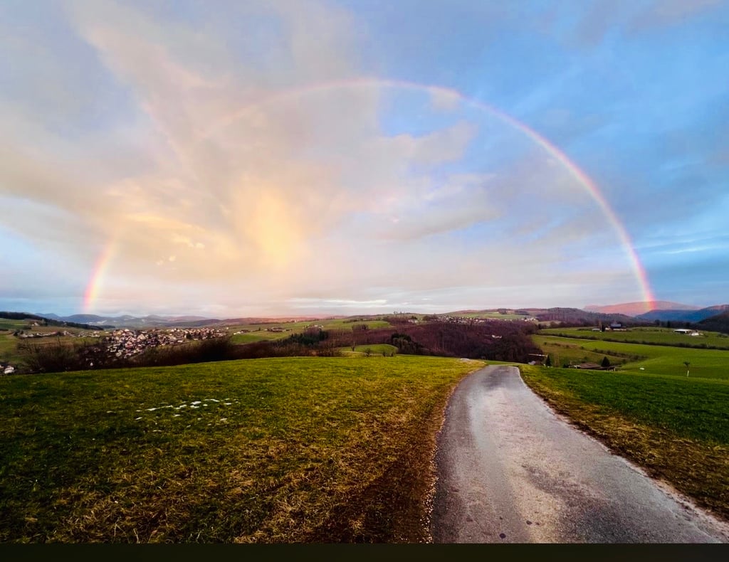 Regenbogen über Feldern und einer Landstrasse in der Abenddämmerung